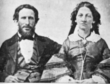 A man and woman, shown from the waist up. He has dark bushy hair and a beard and is wearing a three-piece suit with wade lapels and a bow tie. She has dark hair and wears a 19th-century dress with lace collar and bell sleeves.