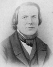 Head and bust of a man with a high forehead, hair reaching his shoulders, wearing a 19th-century three-piece suit and a cravat.